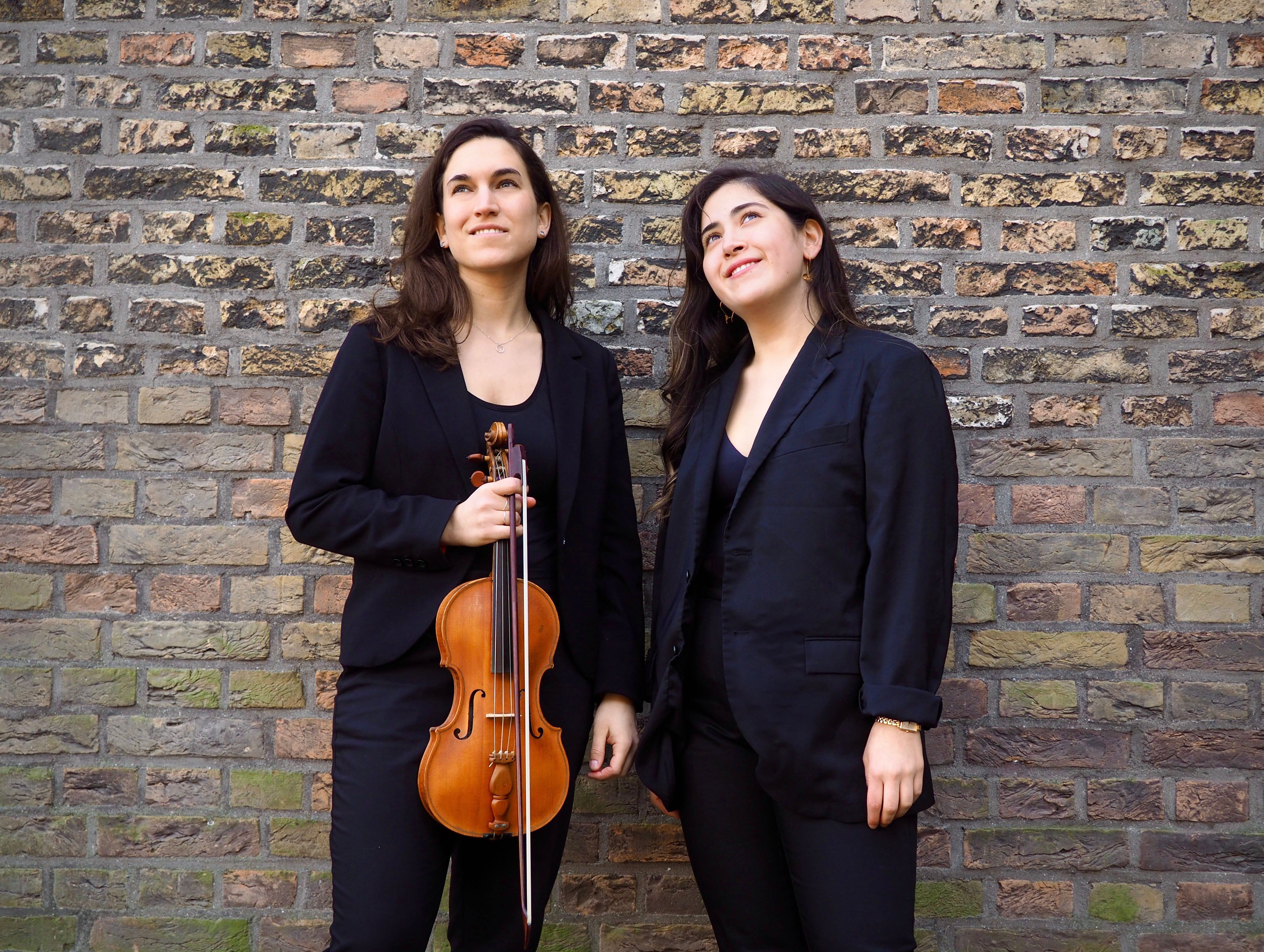 Ai Horton and Elana Cooper with her violin looking up in front of a brick wall.